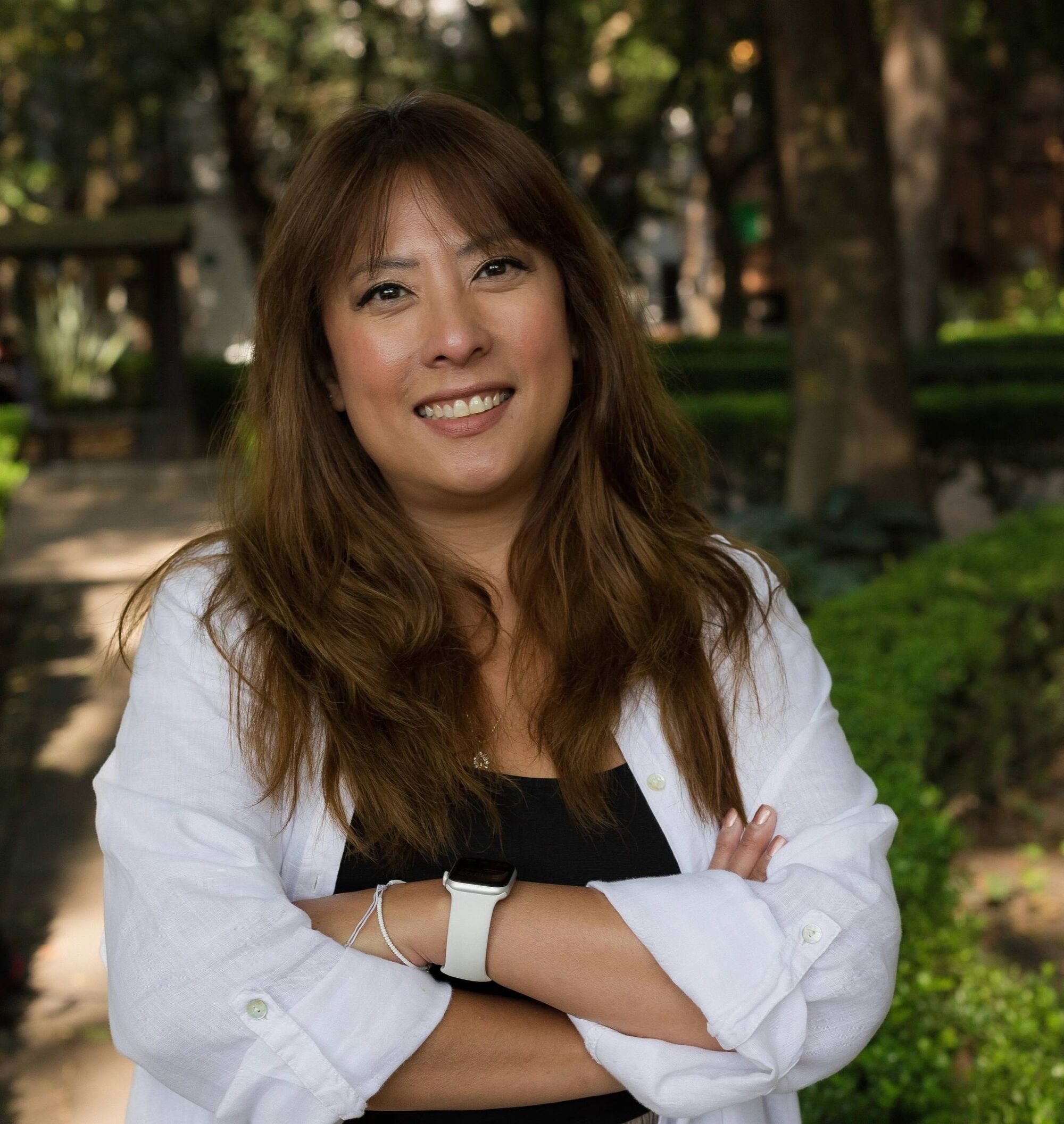 A woman with long brown hair, brown eyes, and an open smile stands with her arms crossed in a white shirt in front of trees.