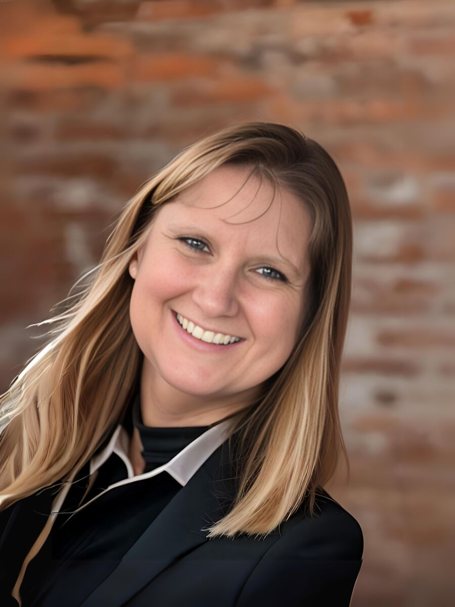 A woman with light brown/reddish hair, brown eyes, and a bright smile. She is wearing a black necklace and blue jacket.