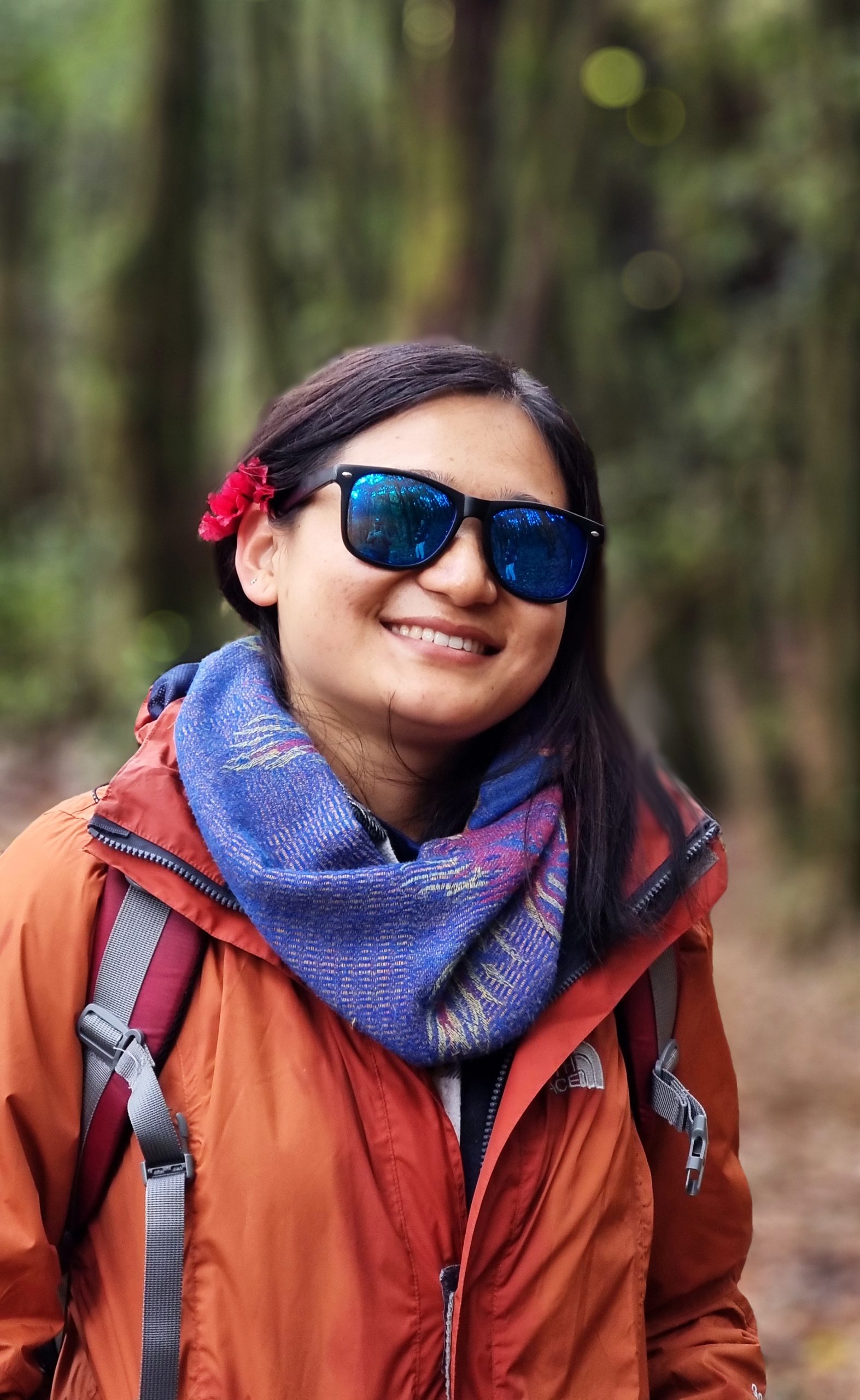 A woman with long dark hair with a red flower tucked behind the ear, sunglasses, and a bright smile. She is wearing a purple scarf and a orange outdoors jacket.