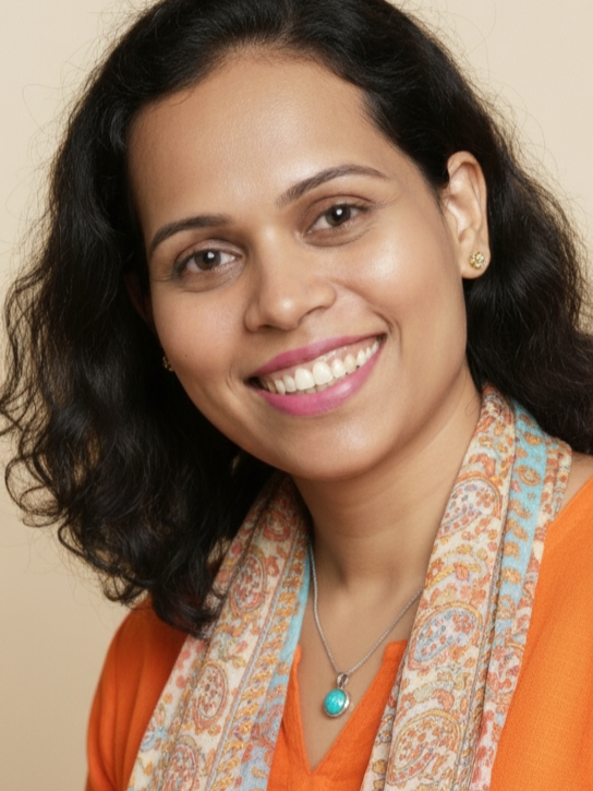 Akshaya Rane - a woman with flowing dark hair, brown eyes, and a wide smile, wearing an orange top, colorful scarf, and blue pendant.