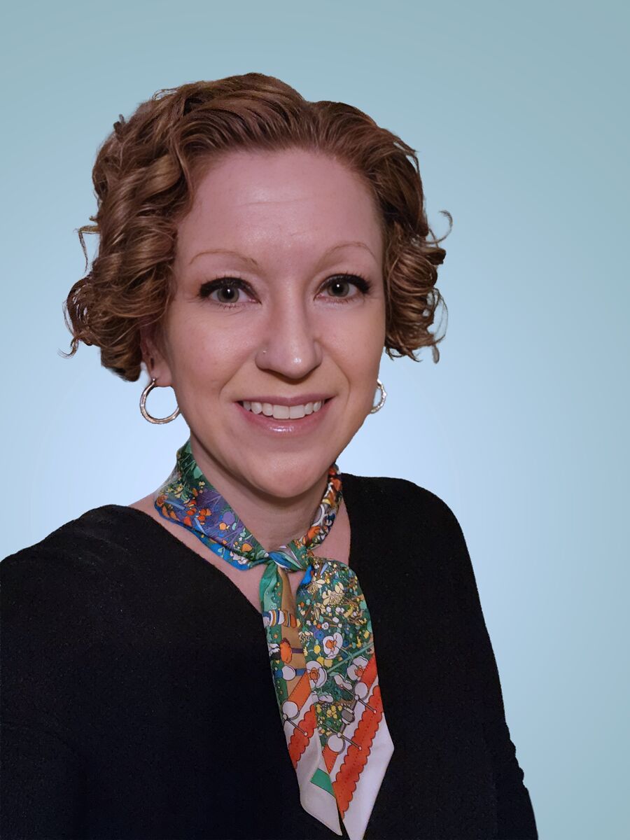 Portrait of Carrie Smaha - woman with short curly red hair, bright smile and wearing a colorful scarf tied around her neck. 