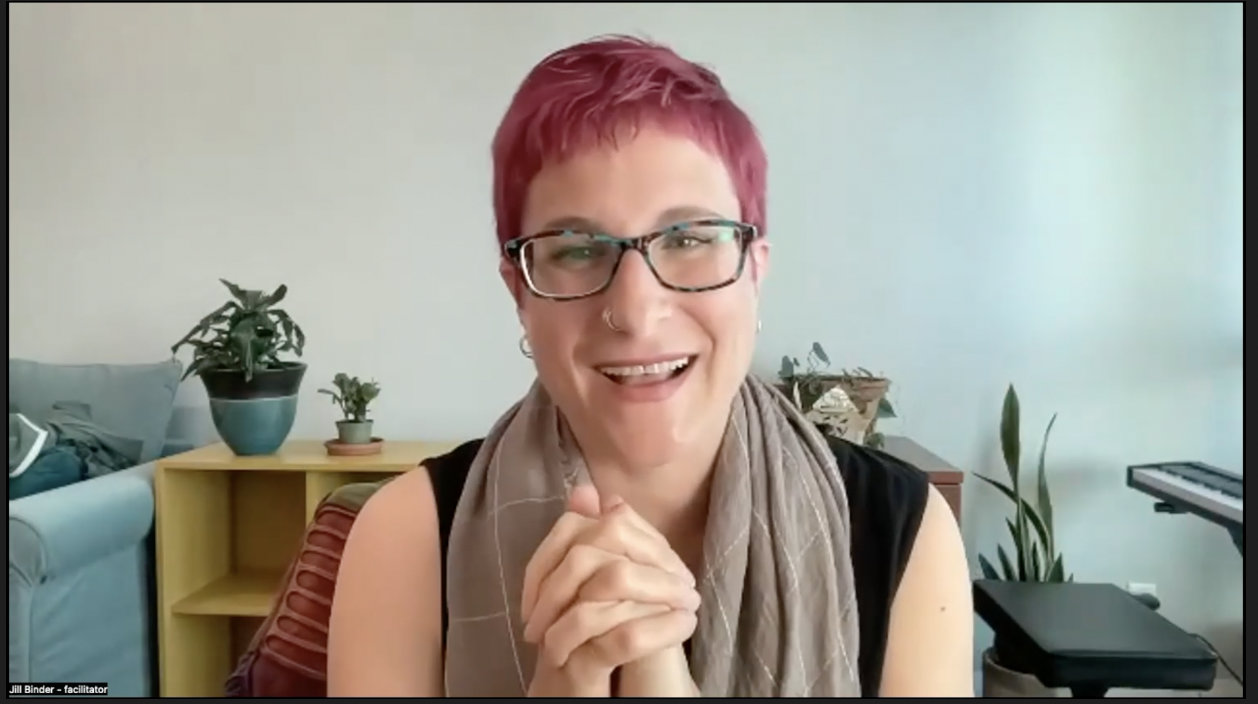 A woman with dark pink hair, glasses, a nose ring, and a ready smile, with hands clasped in front of her and plants,...